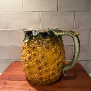 Puede incluir: Una jarra de cerámica con forma de piña con un cuerpo texturizado en amarillo y marrón. El borde superior presenta hojas verdes esculpidas y un asa verde curva se extiende desde el lateral. La superficie tiene un acabado brillante.