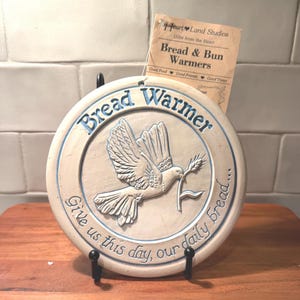 May include: A round, off-white ceramic bread warmer featuring a raised relief of a dove holding a wheat stalk. The text "Bread Warmer" and "Give us this day, our daily bread..." are embossed in blue. A paper tag from Heart Land Studios is attached.