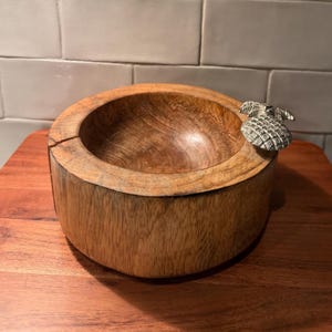 May include: A round, natural-toned wooden bowl with a thick, textured grain and a visible crack on the upper rim. A small, metallic, silver-colored pineapple-shaped ornament is attached to the outer edge of the bowl's rim, adding a distinct detail.