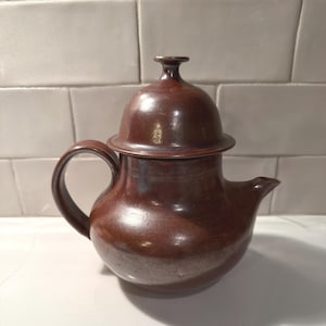 Pode incluir: Uma chaleira de cerâmica marrom feita à mão com tampa e alça curva. A chaleira tem um bico para servir e um corpo arredondado. A tampa tem um pequeno botão na parte superior. A chaleira é provavelmente usada para preparar chá.