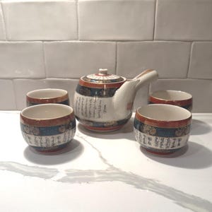 May include: A ceramic tea set consisting of a side-handled teapot and three matching cups. The pieces feature a white base with bands of dark blue and orange, decorated with gold floral patterns and black Japanese calligraphy script throughout.