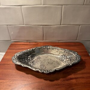 May include: An oval-shaped silver-toned metal tray featuring an ornate embossed floral pattern along the scalloped edges. The interior surface of the tray shows signs of wear with visible tarnishing and a weathered, textured metallic finish.