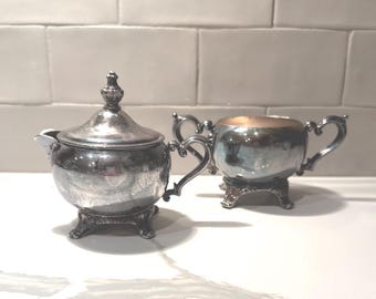 Juego antiguo de jarrita para crema y azucarero de plata con base y tapa, asas ornamentadas en forma de volutas, 5" y 6" de ancho.