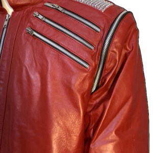 May include: A bright red leather jacket featuring a stand-up collar with a zipper, two diagonal zippered chest pockets, and a silver studded panel on the shoulder. The jacket includes a long vertical zipper along the sleeve and a zippered pocket.