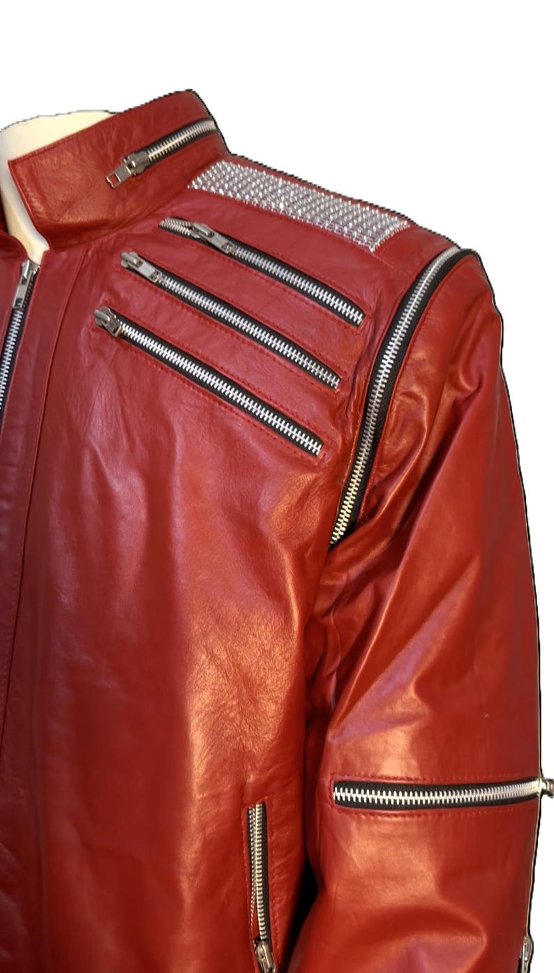 May include: A bright red leather jacket featuring a stand-up collar with a zipper, two diagonal zippered chest pockets, and a silver studded panel on the shoulder. The jacket includes a long vertical zipper along the sleeve and a zippered pocket.
