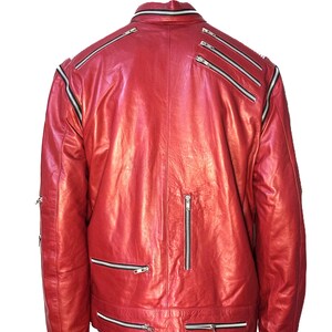 May include: A bright red leather jacket featuring multiple silver-toned metal zippers across the back, shoulders, and lower hem. The jacket has a high collar with a zipper detail and long sleeves, showcasing a structured design with visible seams.