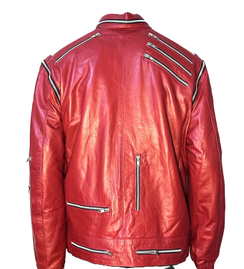 May include: A bright red leather jacket featuring multiple silver-toned metal zippers across the back, shoulders, and lower hem. The jacket has a high collar with a zipper detail and long sleeves, showcasing a structured design with visible seams.