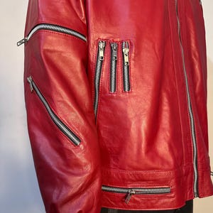 May include: A bright red jacket featuring multiple silver-toned metal zippers. The garment includes a long vertical front zipper, two diagonal zippers on the sleeve, three vertical chest zippers, and a horizontal zipper along the lower hem.