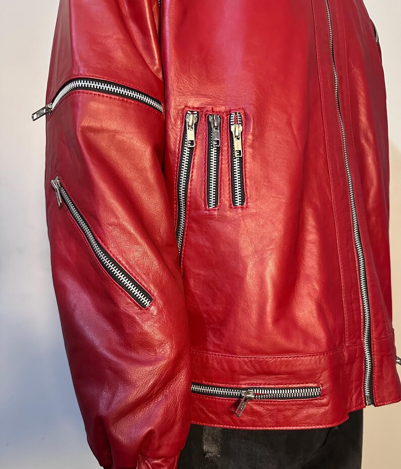 May include: A bright red jacket featuring multiple silver-toned metal zippers. The garment includes a long vertical front zipper, two diagonal zippers on the sleeve, three vertical chest zippers, and a horizontal zipper along the lower hem.