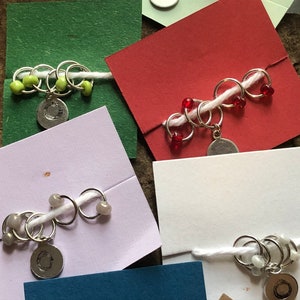 May include: Five sets of stitch markers with different colored beads. Each set has a silver charm with a letter 'C' on it. The stitch markers are on white cards with different colored backgrounds.