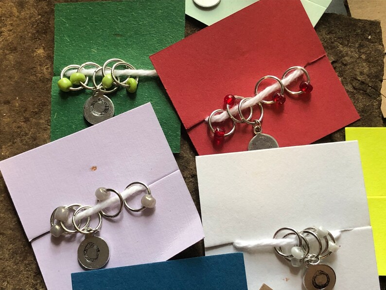 May include: Five sets of stitch markers with different colored beads. Each set has a silver charm with a letter 'C' on it. The stitch markers are on white cards with different colored backgrounds.