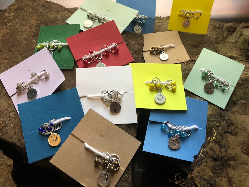 May include: Assortment of colorful stitch markers with beads and charms on white cards. The stitch markers are made of metal and have a circular shape. The beads are various colors, including red, blue, green, and yellow. The charms are silver and have a circular shape with a design.