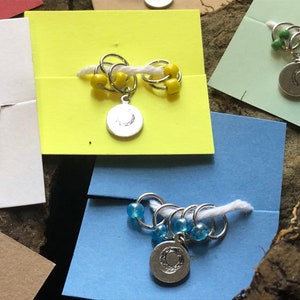 May include: Four sets of stitch markers with different colored beads. Each set has a silver charm with a crescent moon design. The stitch markers are on different colored backgrounds.