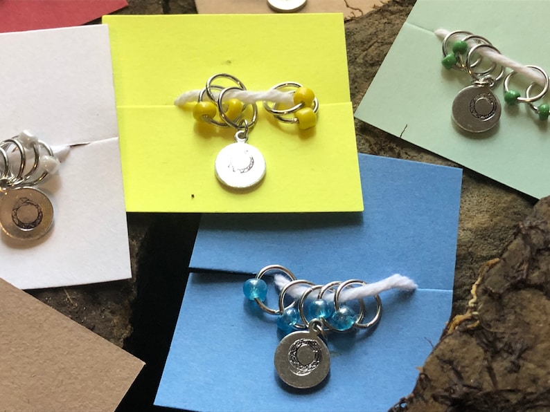 May include: Four sets of stitch markers with different colored beads. Each set has a silver charm with a crescent moon design. The stitch markers are on different colored backgrounds.