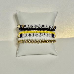 May include: Stack of beaded bracelets with the words "MILWAUKEE" and "BREWERS" spelled out in white letter beads. The bracelets feature black, yellow, gold, and white beads. The bracelets are displayed on a white pillow.