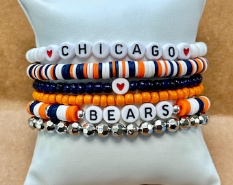May include: A collection of beaded bracelets in white, navy blue, orange, and silver. One bracelet spells out "CHICAGO" with red heart accents, and another says "BEARS". The bracelets are displayed on a white cushion.