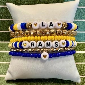 May include: Stack of six bracelets in blue, yellow, gold, and white. The bracelets spell out "LA RAMS" with gold heart accents. The bracelets are displayed on a white pillow.