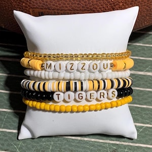 May include: Stack of beaded bracelets in black, white, and gold. One bracelet spells out "MIZZOU" and another spells "TIGERS" in gold letters. The bracelets are displayed on a white pillow with a football in the background.