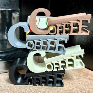 May include: Stacked 3D-printed "COFFEE" signs in various colors: gray, brown, silver, off-white, and black. The letters are thick and blocky, creating a layered effect. The signs are arranged on a wooden surface, with a coffee maker visible in the background.
