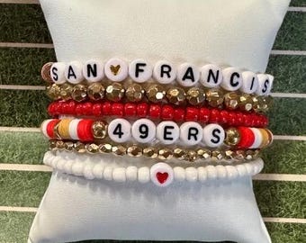 May include: Stack of beaded bracelets in white, gold, and red, with the words "SAN FRANCISCO" and "49ERS" spelled out in white letter beads. One bracelet features a heart-shaped bead. The bracelets are displayed on a white pillow.