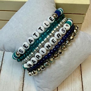 May include: Stack of beaded bracelets in shades of blue, teal, silver, and gold. One bracelet spells out "SEATTLE" and another "MARINERS" in white letter beads. The bracelets are displayed on a white pillow.