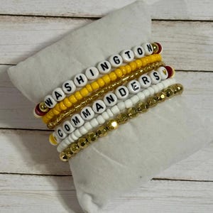 May include: Stack of beaded bracelets in gold, yellow, white, and red. The bracelets spell out "WASHINGTON COMMANDERS" with white letter beads. The bracelets are displayed on a white pillow.
