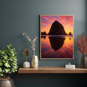 May include: A framed landscape photograph of a coastal scene at sunset. The image features a large rock formation silhouetted against a vibrant sky of orange, pink, and purple hues. The water reflects the sky, creating a mirror image.
