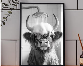 May include: Black and white framed print of a Highland cow taking a shower. The cow is covered in soap suds under a silver shower head. The print is displayed on a wooden surface with a white tiled background.