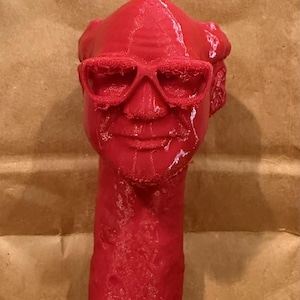 May include: A solid red sculpture of a face wearing sunglasses. The sculpture has a neck and a head with hair. The face has a mustache and a smile. The sculpture is made of a smooth material.