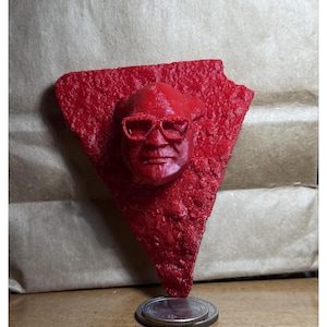 May include: A red, textured sculpture of a face wearing glasses, mounted on a triangular base. The sculpture is placed on top of a stack of coins. The background is a brown paper.
