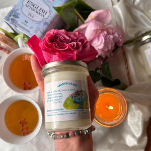 Peut inclure: Une bougie en pot de verre transparent avec une étiquette blanche portant le texte KRASNYLLA.ART, Love is patient, love is kind, It does not envy, It does not boast, It is not proud, 1 Kor 13:4, TEA IN THE GARDEN, et HANDCRAFTED - NATURAL SOY CANDLES.