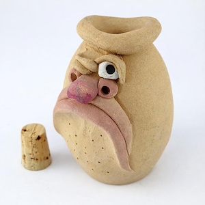 Op de afbeelding: Een bruin keramieken fles met een gebeeldhouwd gezicht, inclusief een grote neus, één oog en een neerhangende mond. Een kurkstopper staat naast de fles. De fles heeft een uniek, expressief ontwerp.