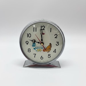 May include: A vintage silver-toned alarm clock with a cream-colored face. The clock features black numerals, black hands, and a red second hand. A colorful illustration of a hen and chicks is on the face. The clock sits on a silver base.