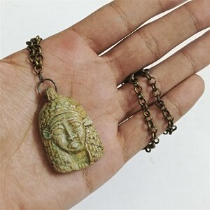 Może przedstawiać: Naszyjnik z wisiorkiem w stylu vintage z rzeźbioną twarzą i nakryciem głowy. Wisiorek jest w kolorze jasnozielonym ze skomplikowanymi detalami, zawieszony na łańcuszku w kolorze brązu. Naszyjnik jest trzymany w dłoni.