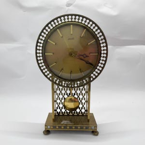 May include: A vintage brass-toned mantel clock with a round face and a decorative openwork frame. The clock features a pendulum and a rectangular base with small feet. The clock face has gold-colored hands and hour markers.