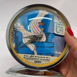 May include: A vintage, round, chrome-plated alarm clock. The clock face features an illustration of a person in striped pajamas dreaming of a professional wrestler. The text on the clock face reads "The girl dreamed of a Professional wrestler..."