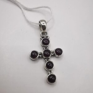 May include: A silver-toned pendant in the shape of a cross, featuring six round, dark red gemstones. The pendant is suspended from a thin, white cord, and is set against a white background.