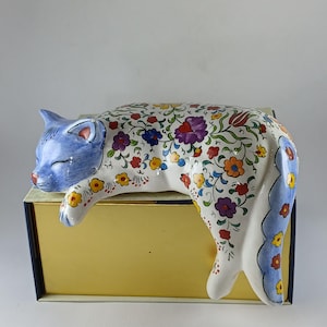 Puede incluir: Figura de gato de cerámica, durmiendo sobre una caja dorada. El gato es blanco con cabeza y cola azules, y está decorado con coloridos estampados florales. Los ojos del gato están cerrados y parece estar descansando.