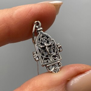 Pode incluir: Um pequeno e ornamentado pingente de prata com um crucifixo dentro de uma moldura retangular. O pingente tem intrincados trabalhos de rolagem e um design articulado. A argola é presa na parte superior e o pingente é segurado entre os dedos.