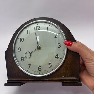 May include: A vintage, wooden-framed clock with a circular face. The clock face is white with black numerals and hands. The clock is marked "SMITHS" and "MADE IN GT BRITAIN". The clock is a dark brown color.