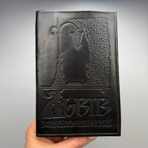 May include: A rectangular black leather journal cover featuring an embossed design of a street lamp and dense foliage. The word "ALBIB" is embossed in a stylized font at the bottom center, with decorative border patterns along the edges.
