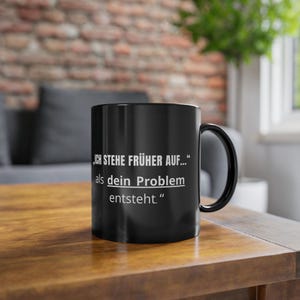 Peut inclure: Mug en céramique noire avec une anse incurvée. Le mug présente un texte blanc qui se lit : "ICH STEHE FRÜHER AUF..." als dein Problem entsteht." Le mug est posé sur une surface en bois.