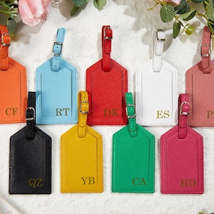 Könnte beinhalten: Eine Sammlung farbiger Gepäckanhänger in Orange, Hellblau, Rot, Weiß, Schwarz, Gelb, Grün und Pink. Jeder Anhänger hat einen Schnallenriemen und einen goldenen Buchstaben. Die Anhänger sind auf einer weißen Oberfläche angeordnet.