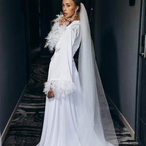 May include: A white wedding dress with long sleeves and feather trim. The dress features a flowing skirt and a long, sheer veil. The bride is standing in a hallway with a patterned carpet.