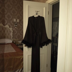 May include: A long, black robe with wide sleeves and a tie waist hangs on a door. The sleeves are trimmed with black feathers. The robe is hanging in a room with a bed and patterned wallpaper.
