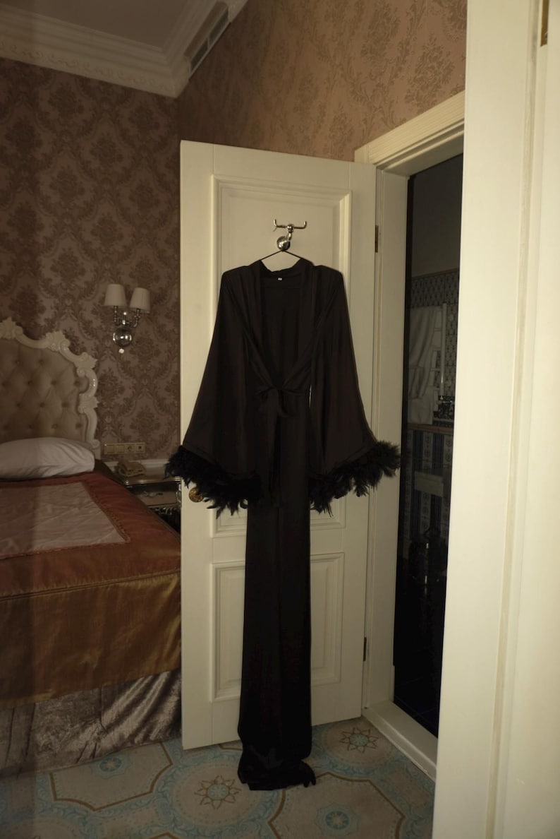 May include: A long, black robe with wide sleeves and a tie waist hangs on a door. The sleeves are trimmed with black feathers. The robe is hanging in a room with a bed and patterned wallpaper.