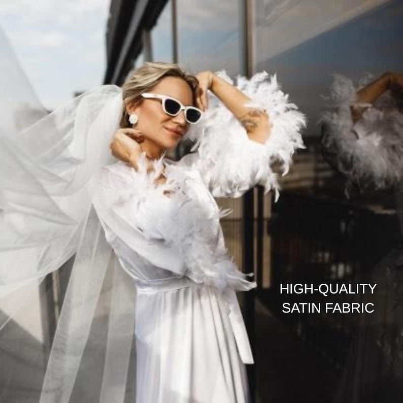 May include: A white satin robe with feather trim and a matching veil. The robe has a tie waist and is paired with white sunglasses. The image includes the text "HIGH-QUALITY SATIN FABRIC".