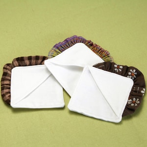 May include: Three white, square cloth pads with folded corners are surrounded by decorative, ruffled edges. The edges feature brown fabric with floral and striped patterns. The pads are likely reusable makeup remover pads.