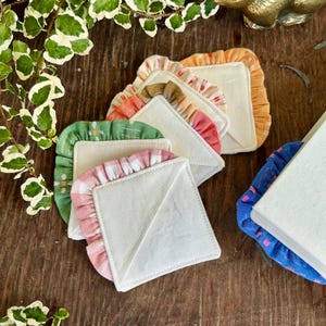 May include: A collection of colorful, square fabric coasters with ruffled edges. The coasters feature various patterns, including gingham and floral designs, in shades of pink, green, orange, and brown. They are arranged on a dark wooden surface.