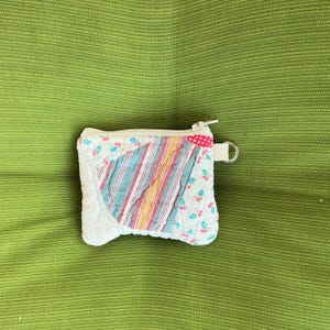 May include: A small, quilted coin purse with a zipper closure. The purse features a patchwork design with a central section of blue, red, and orange stripes. The edges are white with a floral pattern. A small red polka dot accent is on the side.